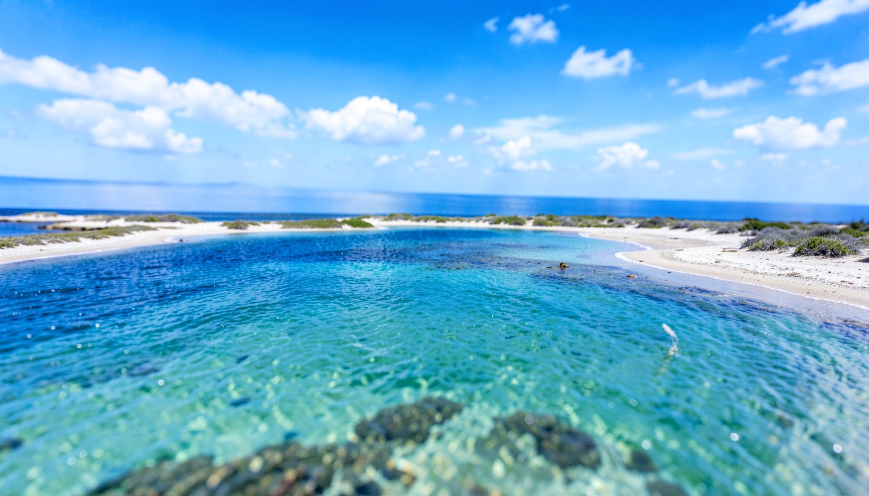 Wide aerial view of a windswept coastal lagoon with turquoise shallow water and a sandy shoreline, under a bright blue sky with scattered white clouds